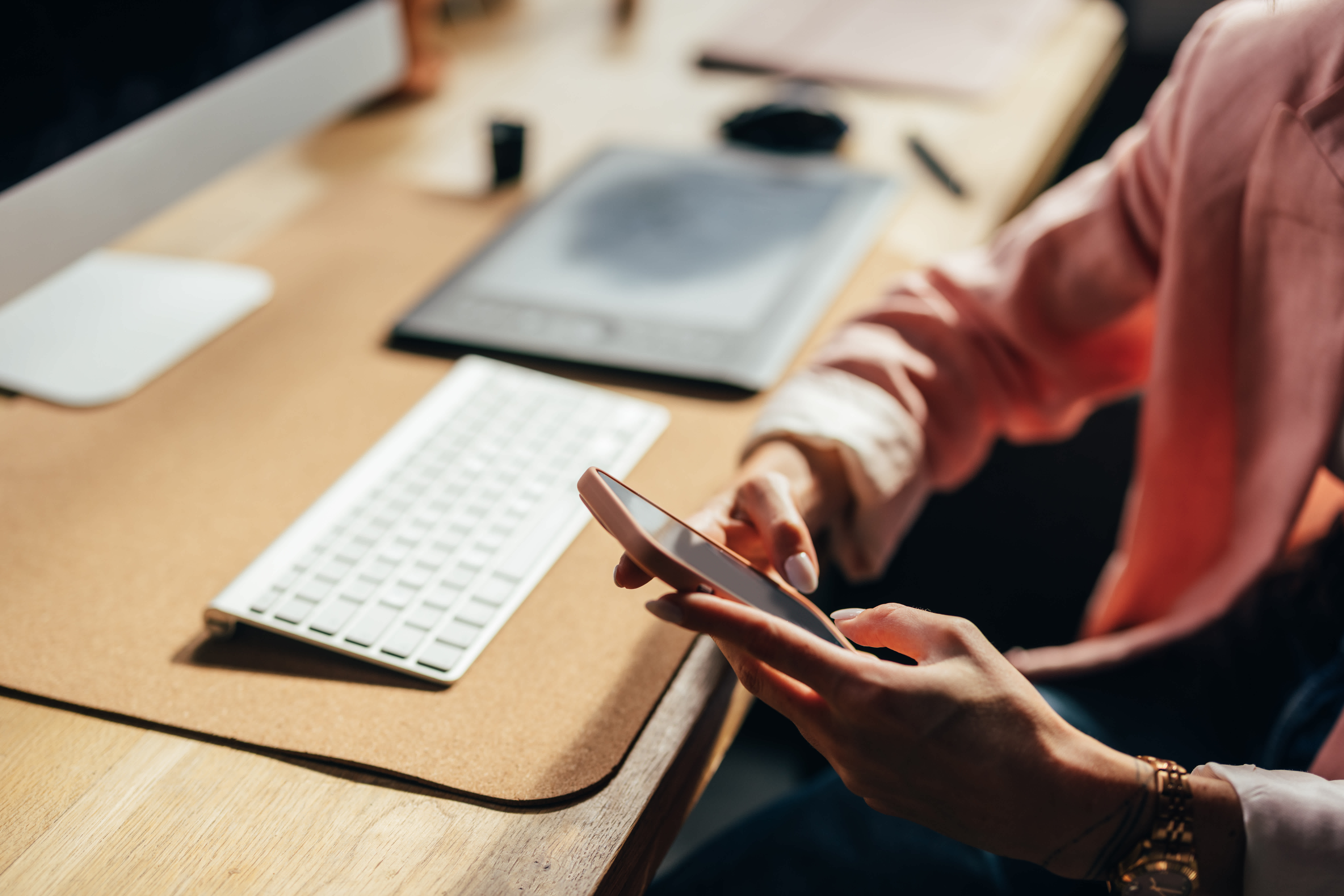 A person in a pink blazer uses a smartphone at a desk with a computer keyboard, tablet, and notepad, suggesting a modern workspace environment.