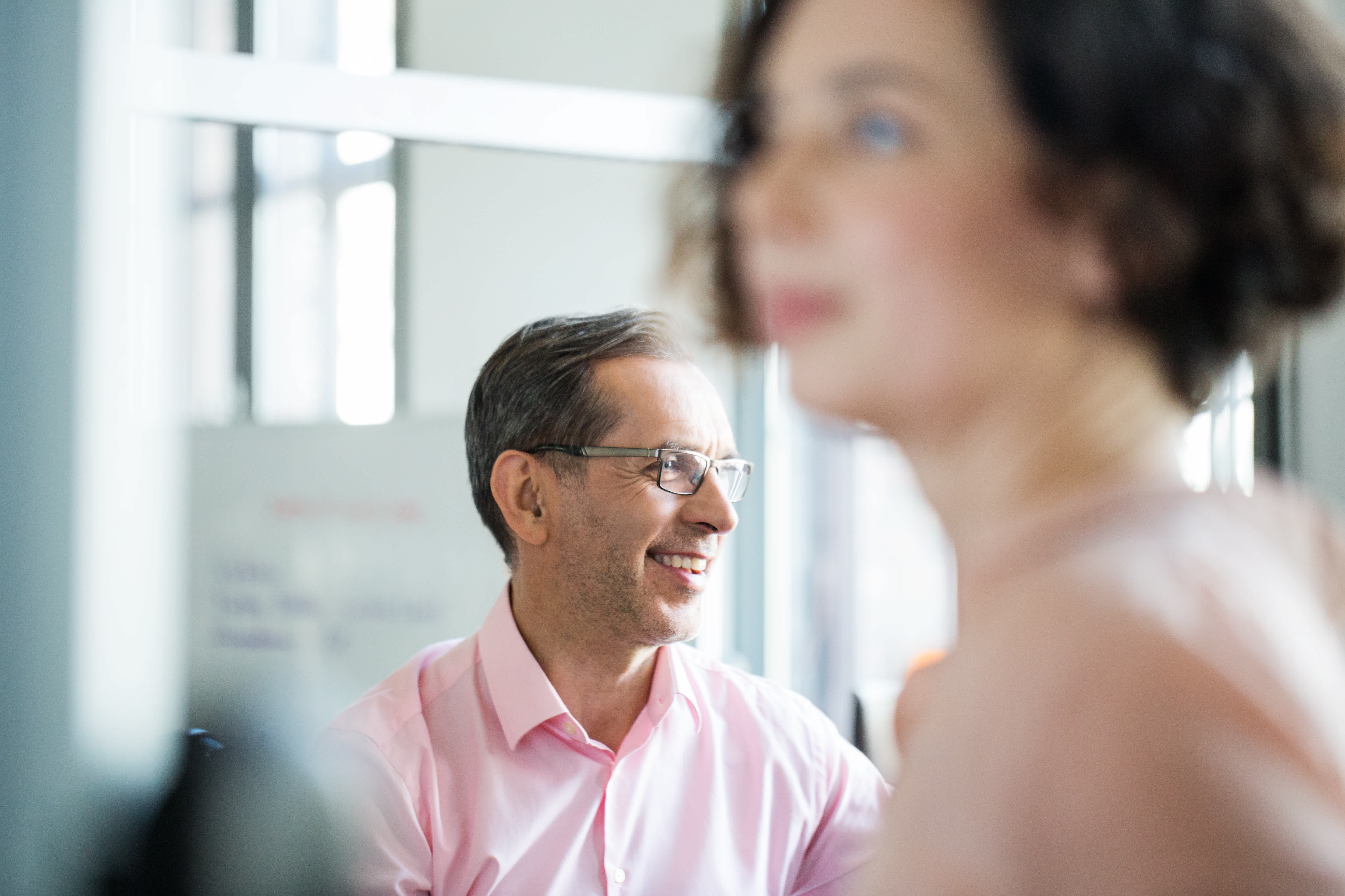 A man in glasses and a pink shirt smiles while sitting indoors; a woman with short dark hair, also in pink, is out of focus in the foreground.