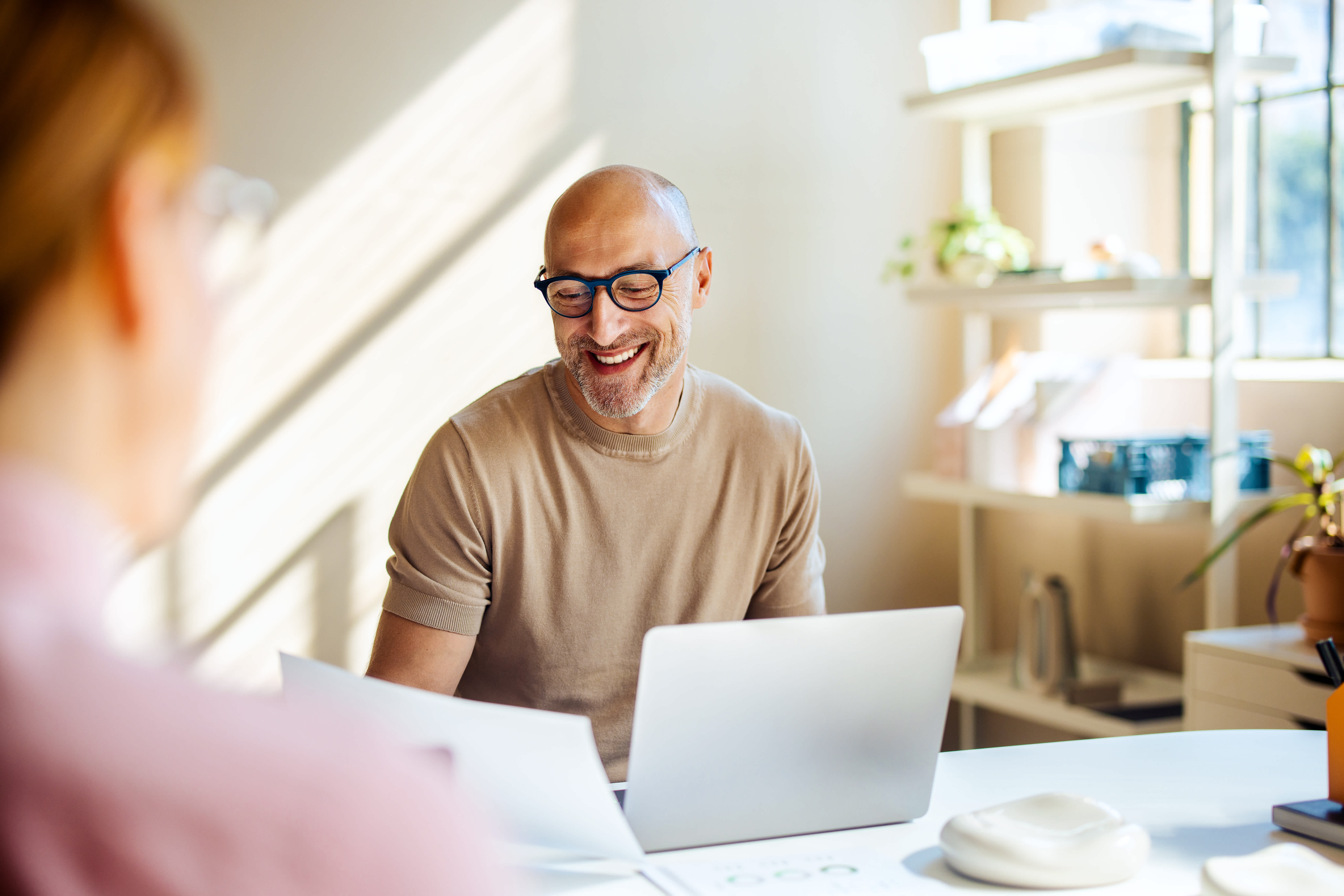 A person with long dark hair and a beard is smiling while working on a laptop. They are wearing an orange sweater and are seated at a desk with papers. The background is blurred with dark tones and lights.