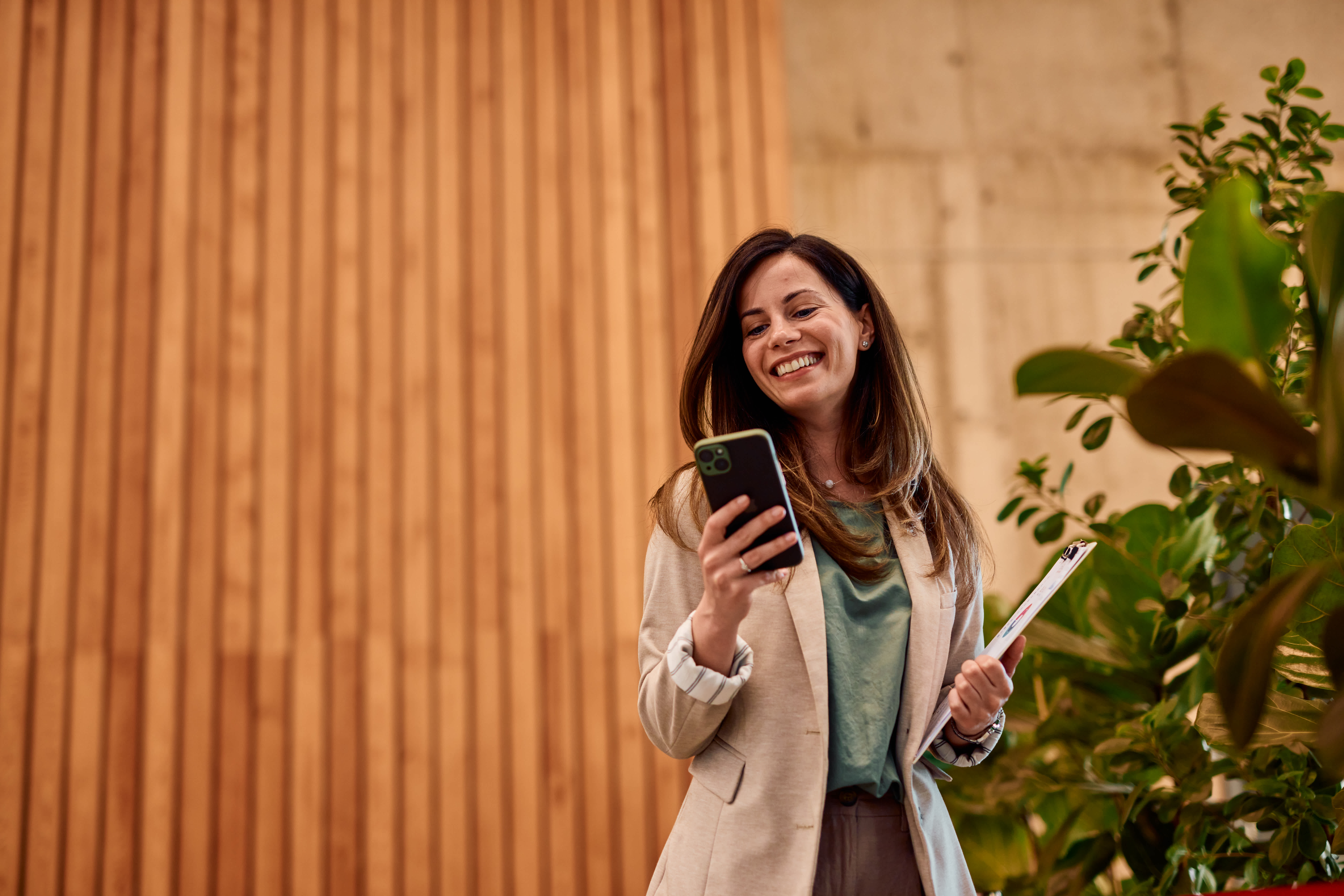 A woman wearing glasses and a dark shirt stands by a green plant, smiling while holding and looking at an open laptop against a plain teal wall.