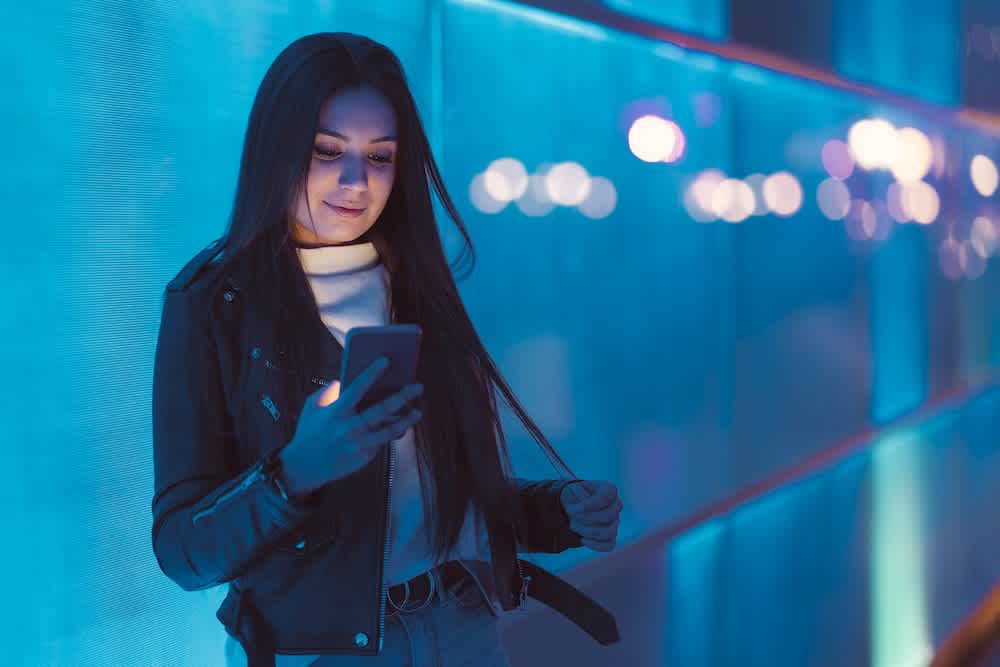 A young woman with long dark hair stands by a blue-lit wall at night, looking at her smartphone and smiling slightly. She is wearing a black leather jacket and a white turtleneck.