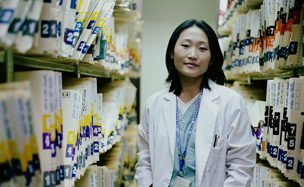 A woman in a white lab coat stands between shelves filled with organized file folders in what appears to be a medical records archive room.
