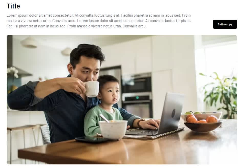 A man holds a toddler and drinks from a cup while working on a laptop at a kitchen table with fruit and notebooks in the background.