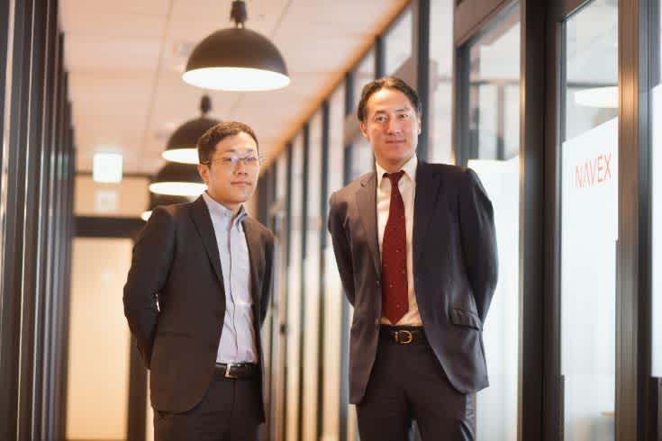 モダンなオフィスのガラス張りの廊下で、スーツを着たビジネスパーソンが並んで立っている様子。