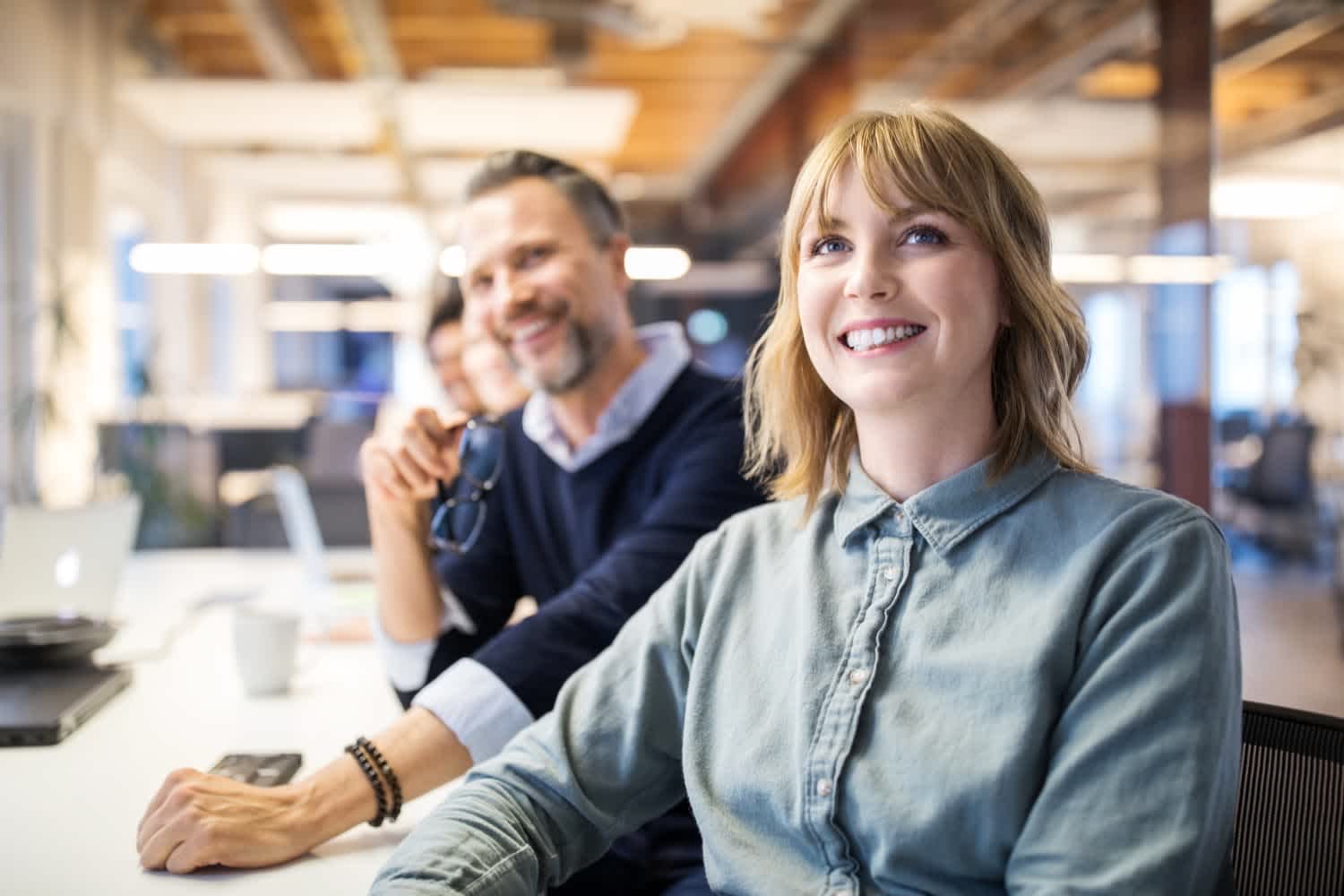 A smiling woman sits at a desk in a modern office, with two colleagues, including a man in glasses, slightly blurred in the background. The setting is bright with exposed beams and computer equipment visible.