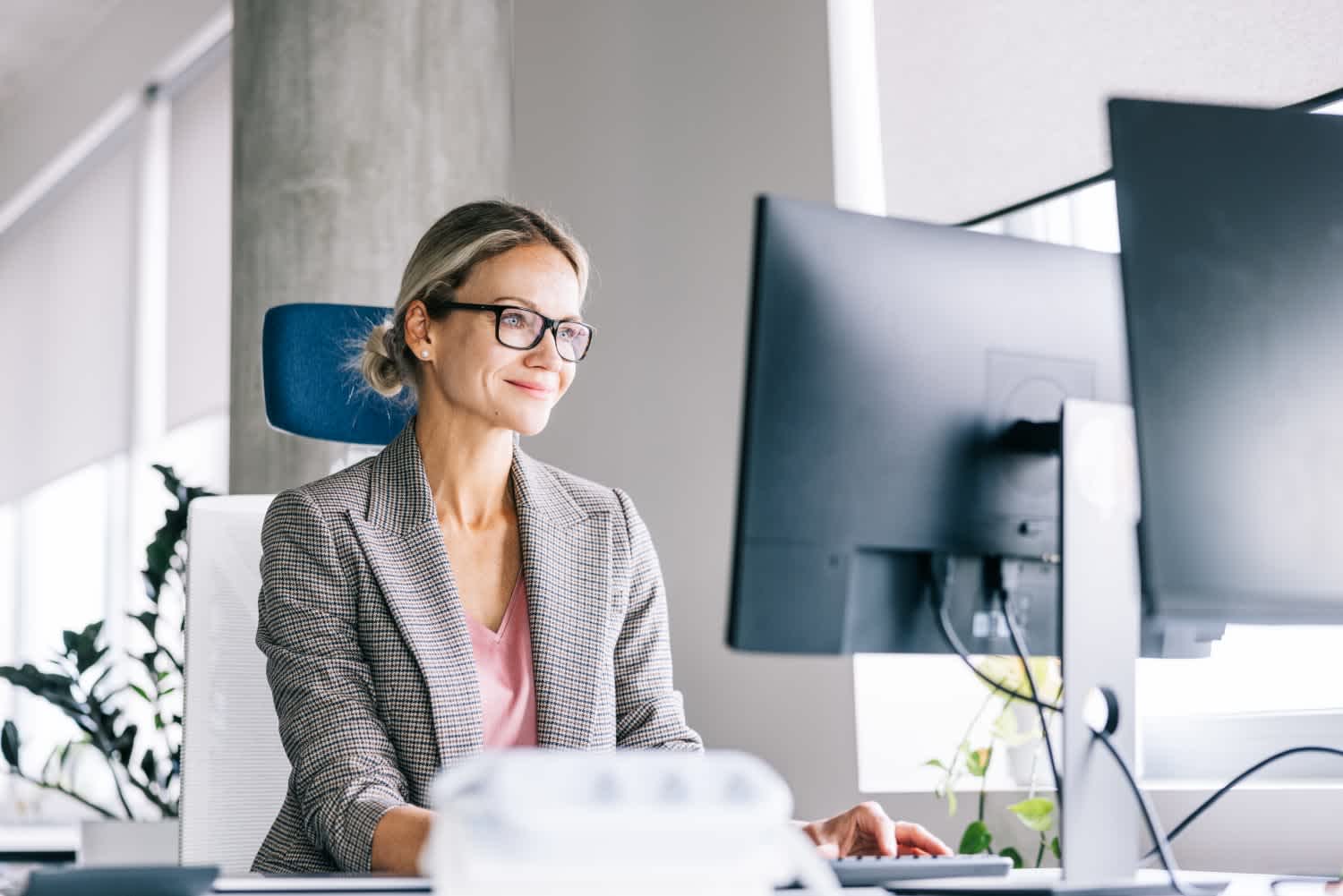A woman wearing glasses and a checked blazer sits at a desk, working on a computer in a bright, modern office with plants in the background.