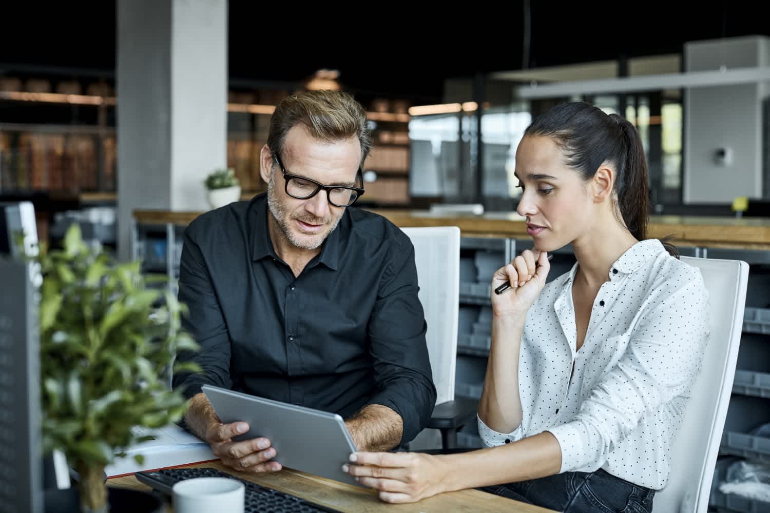 Two colleagues sit at a desk in a modern office, looking at a tablet together. The man wears glasses and a black shirt; the woman, in a white blouse with black polka dots, listens attentively. A plant is visible in the foreground.