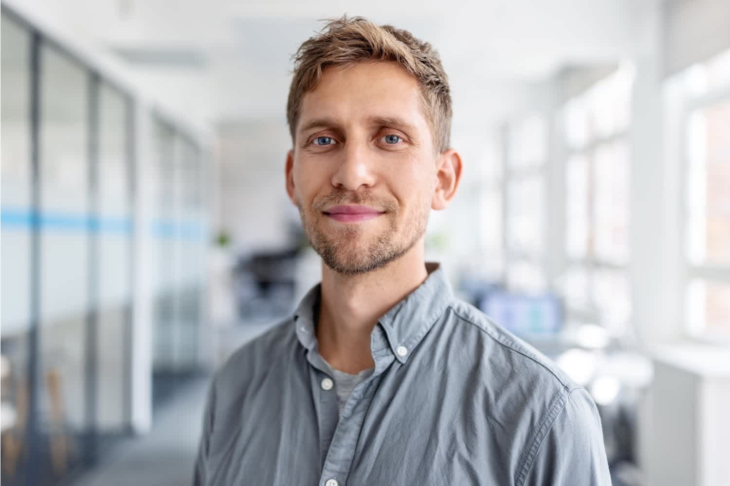 A man with short, light brown hair and a trimmed beard smiles gently, standing in a bright, modern office with glass walls and large windows. He wears a light gray button-up shirt.