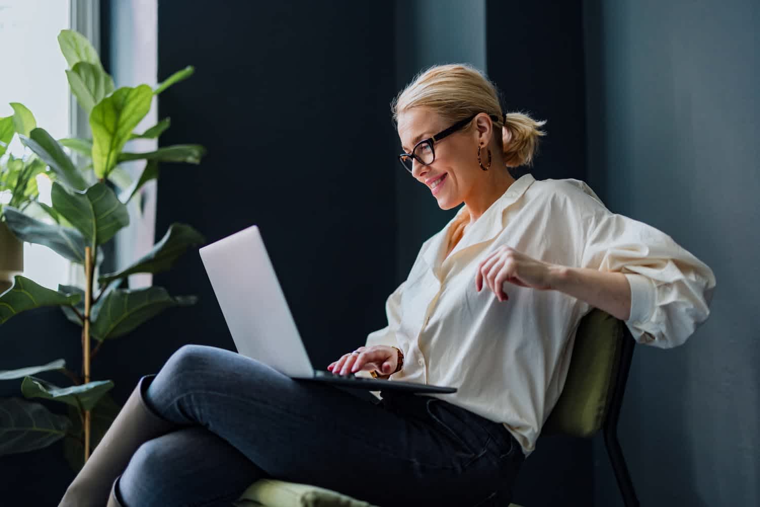 A woman with blonde hair in a bun, wearing glasses, a white shirt, and jeans, sits on a chair smiling while working on a laptop. A green potted plant is next to her by a window.