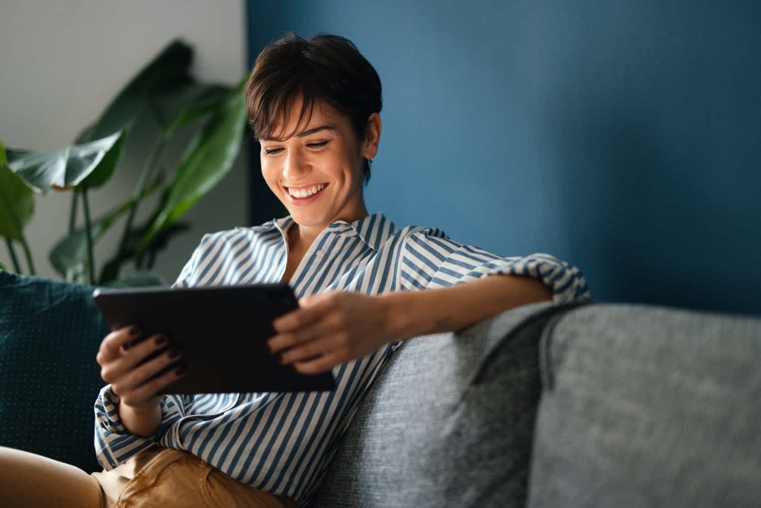 A smiling person with short hair sits on a couch, wearing a striped shirt and holding a tablet, with green plants and a blue wall in the background.