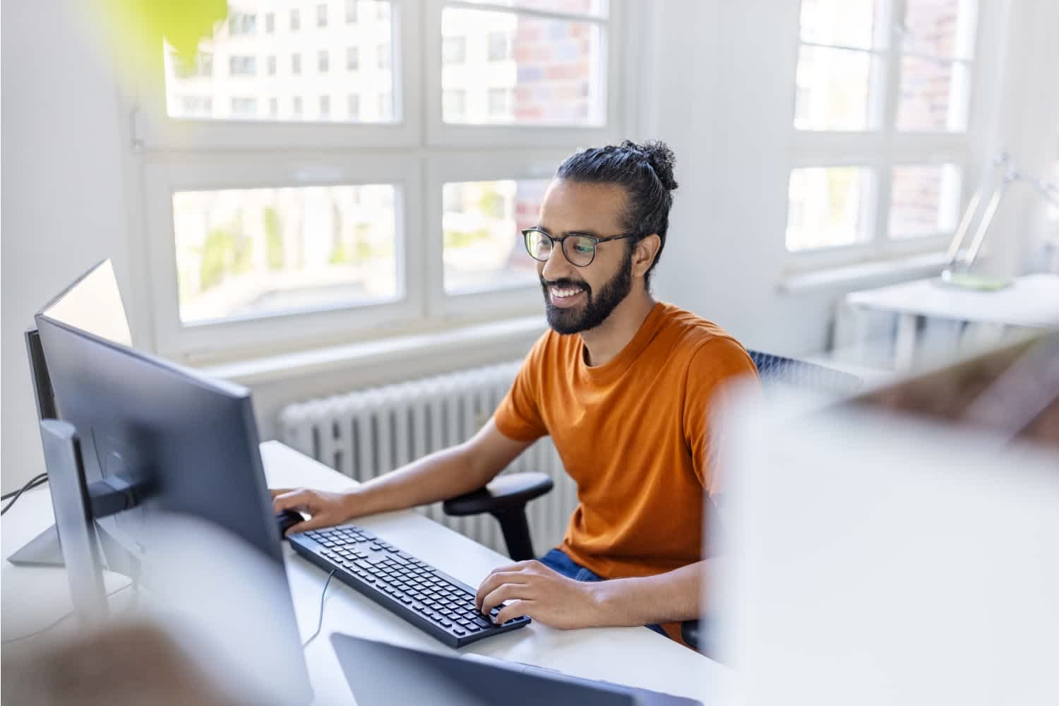 A smiling man with glasses and a beard, wearing an orange t-shirt, sits at a desk working on a computer in a bright, modern office with large windows.