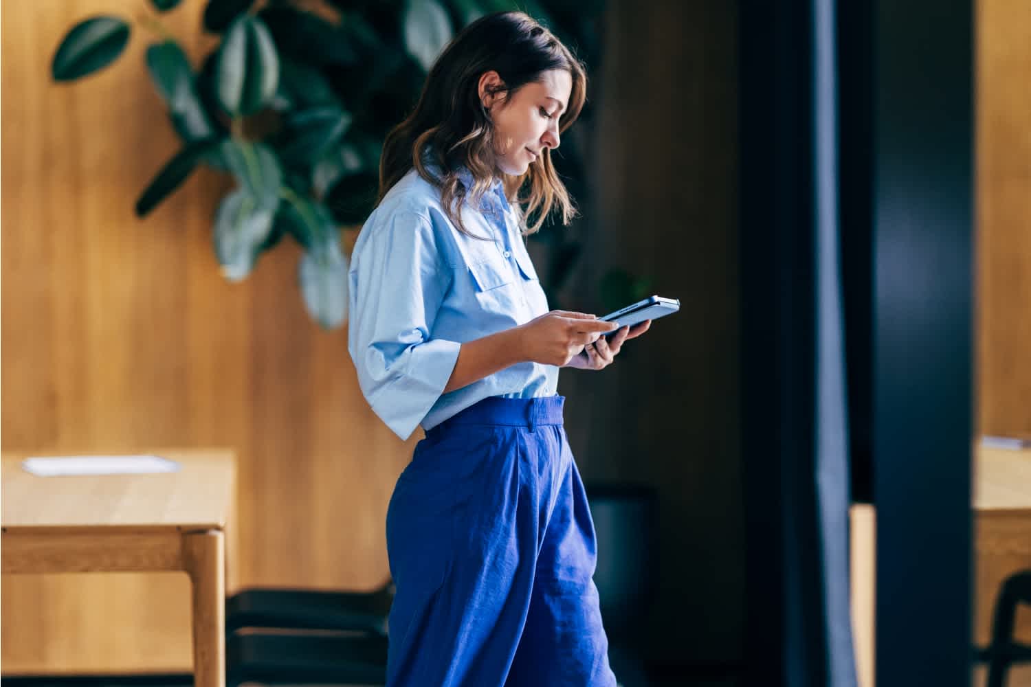 A woman wearing a light blue blouse and blue pants stands indoors, looking down at a tablet in her hands. There is a wooden table and a large leafy plant in the background.