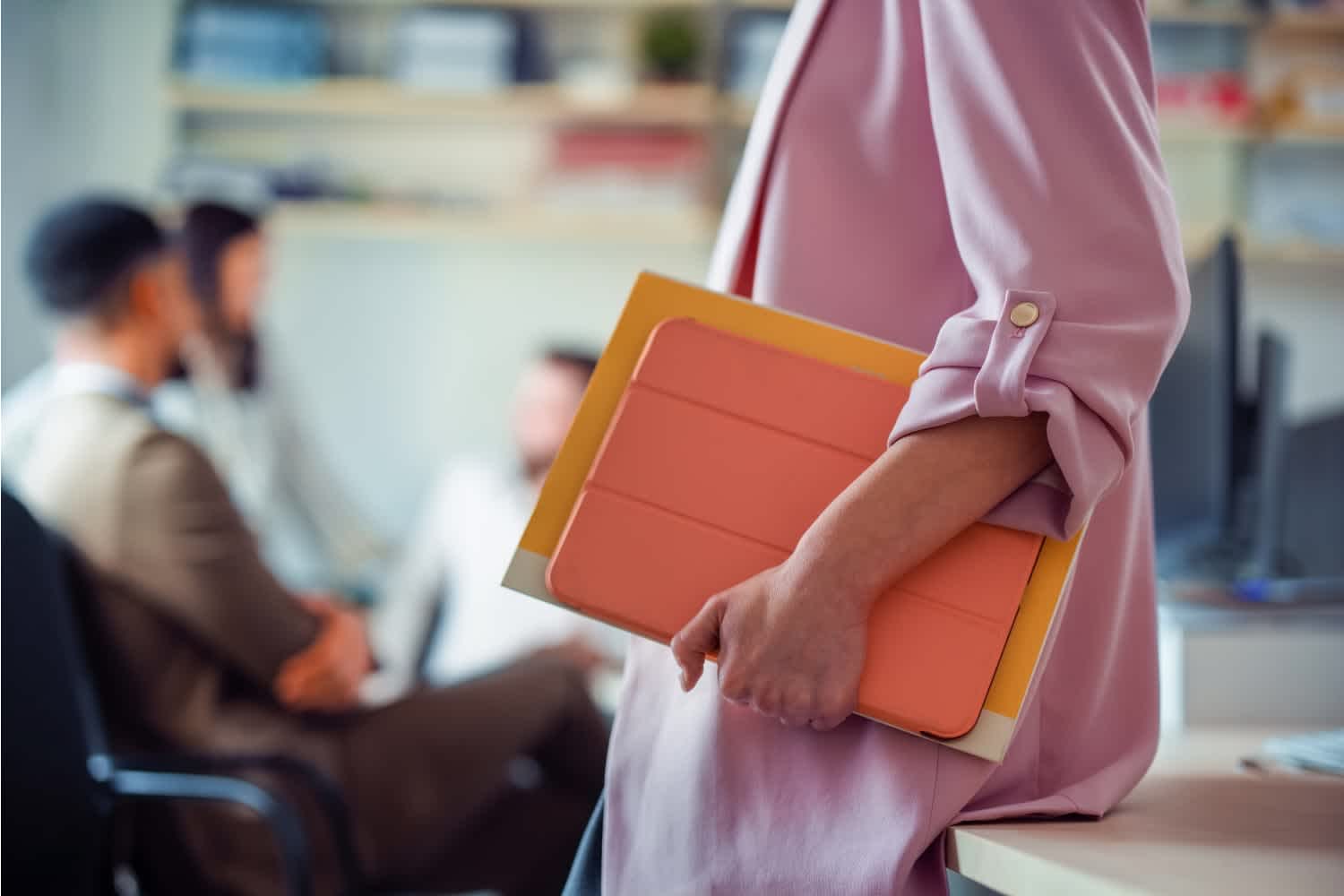 A person in a pink blazer holds folders and a tablet while leaning on a desk. In the background, three blurred people sit and talk in an office setting.