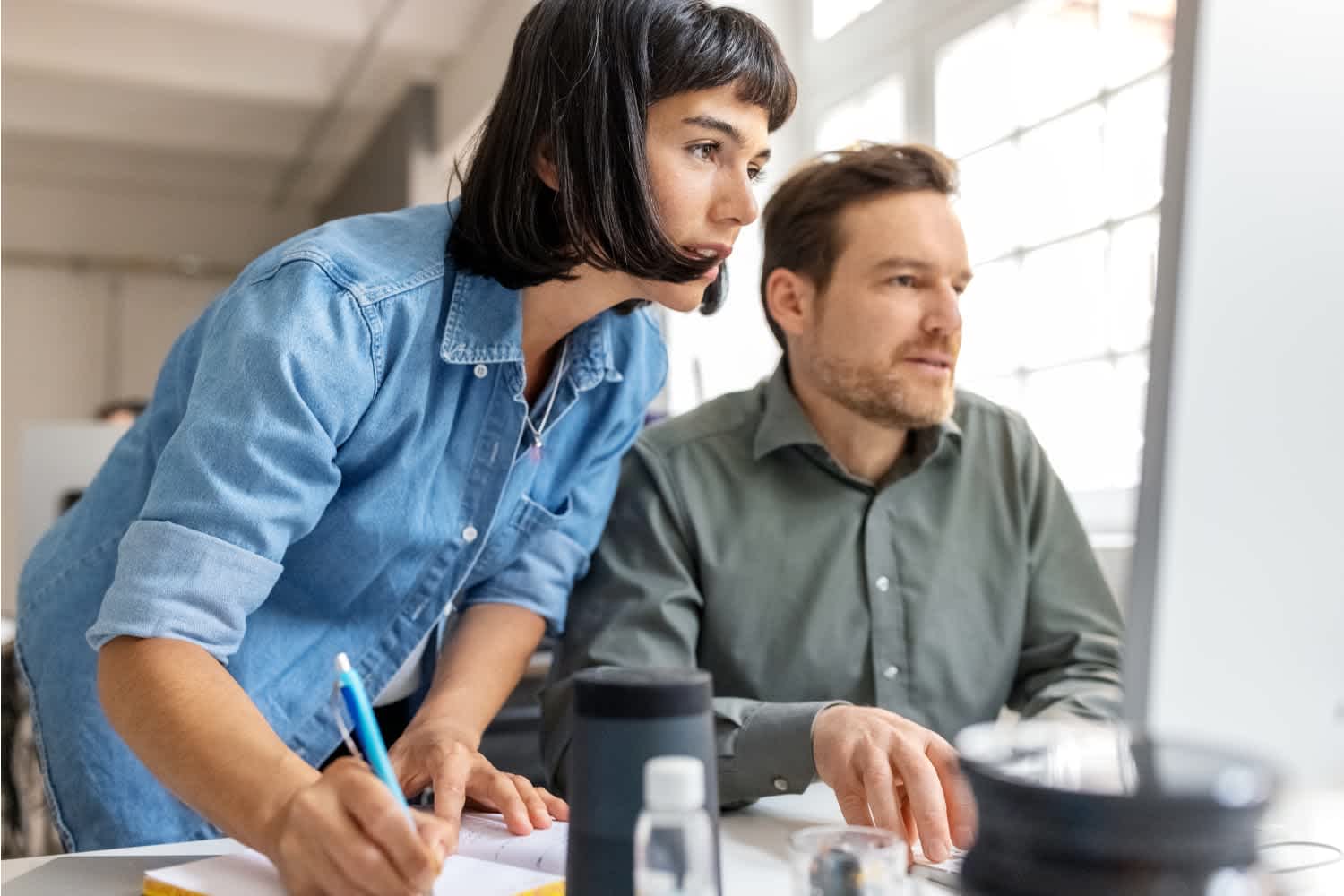 Two people work together at a desk, looking intently at a computer screen. One person is sitting and using the keyboard, while the other stands nearby, holding a pencil and notepad, appearing focused and engaged in discussion.