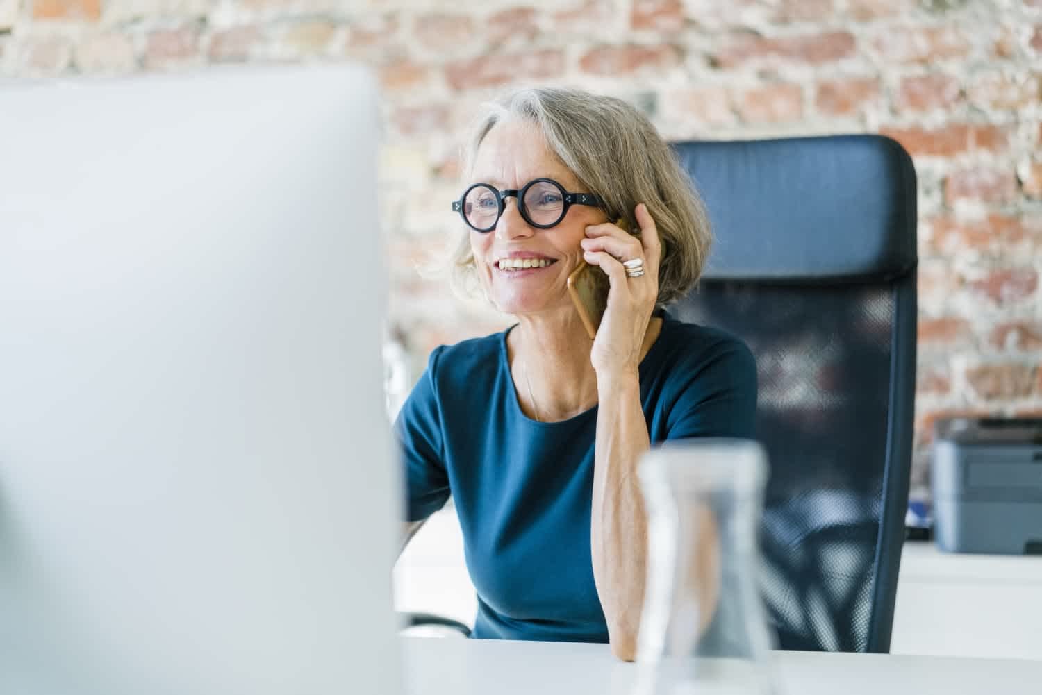 Smiling older woman with gray hair and glasses talks on a smartphone while sitting at a desk with a computer monitor, in a modern office with a brick wall background.