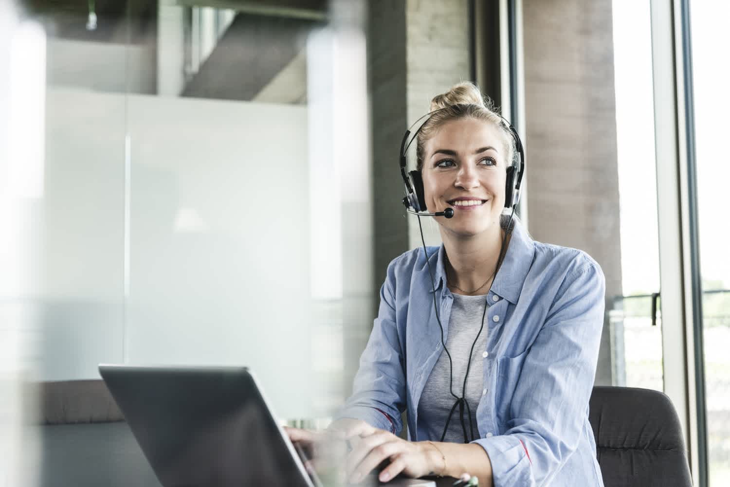 A woman wearing a headset sits at a desk with a laptop, smiling and looking to the side. She appears to be in a modern office setting with large windows and glass walls.