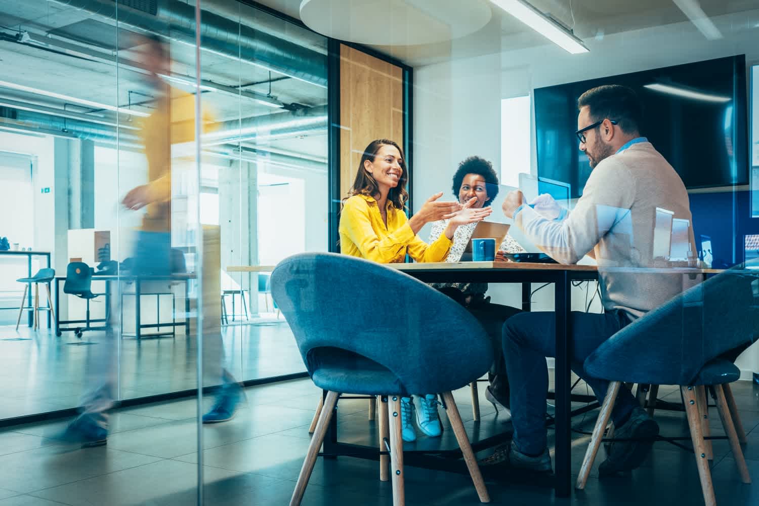 Three people sit at a table in a modern office, engaged in discussion and smiling. One person walks past outside the glass-walled meeting room, creating a blurred motion effect.
