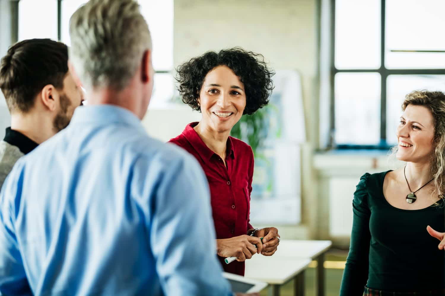 Four people stand together in a bright office, engaged in friendly conversation. A woman in a red shirt smiles warmly at her colleagues, while others listen and smile, creating a positive, collaborative atmosphere.