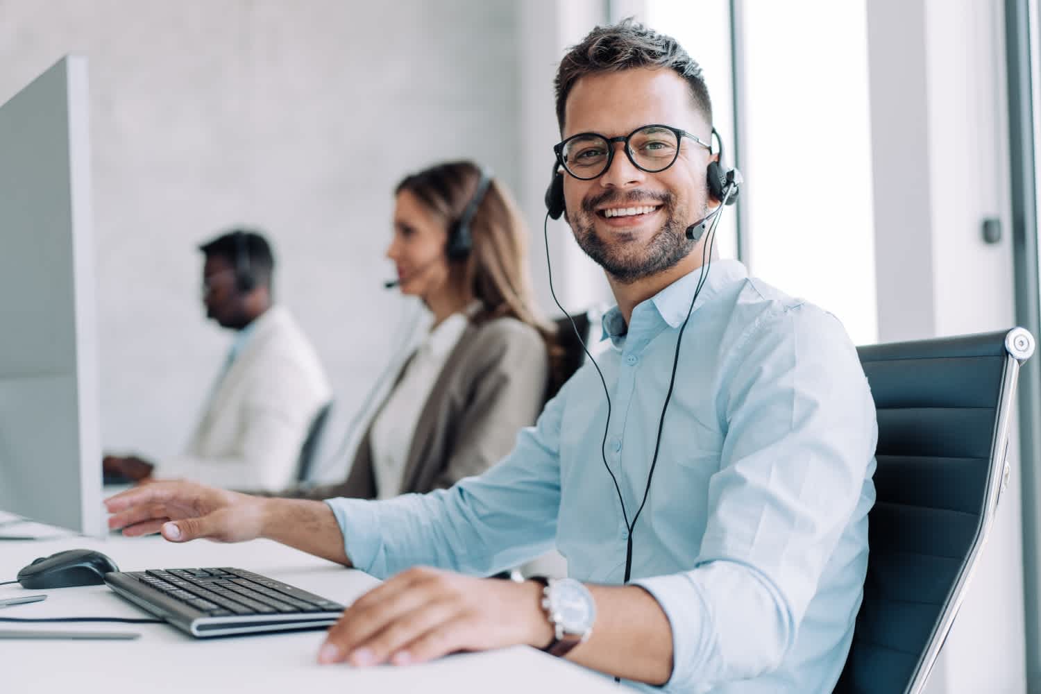 A smiling man wearing glasses and a headset sits at a desk in an office, looking at the camera. Two other people with headsets work at computers in the background.