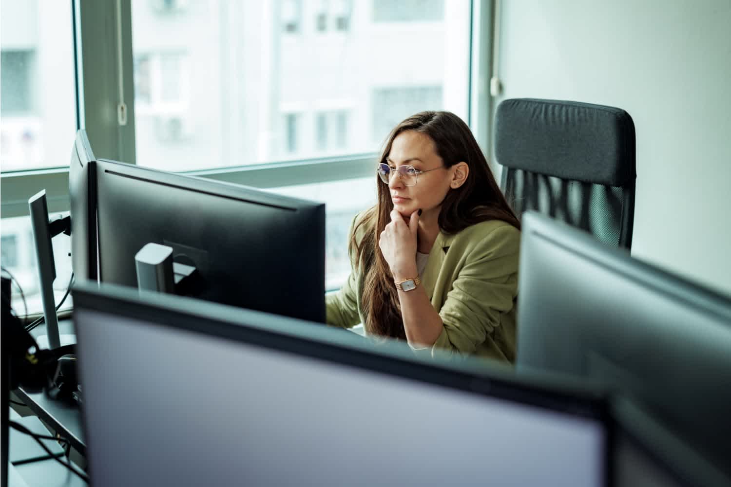 A woman with long brown hair and glasses sits at a desk, looking thoughtfully at a computer monitor in a modern office with large windows.
