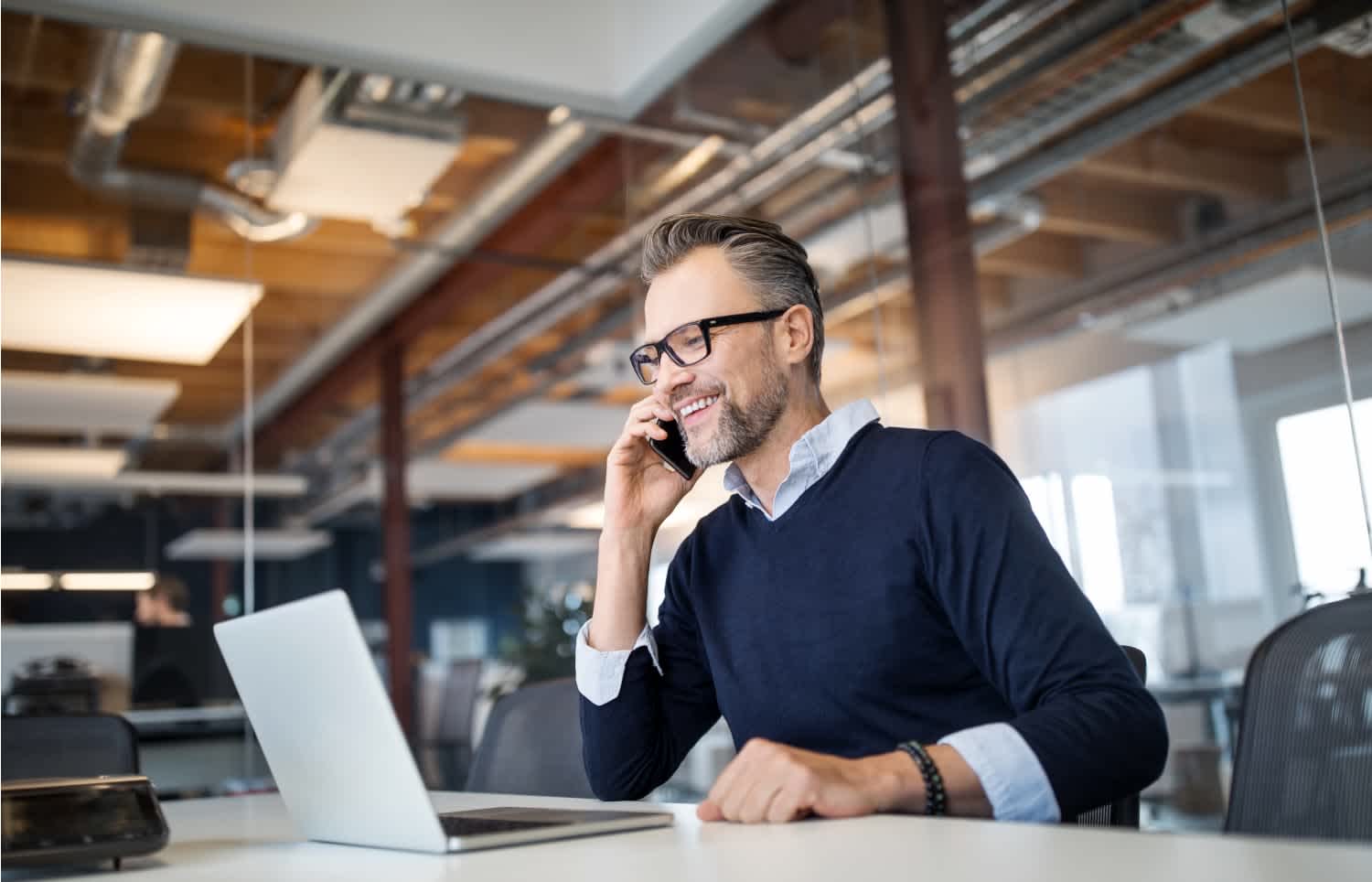 A smiling man wearing glasses and a navy sweater sits at a desk in a modern office, talking on his smartphone and working on a laptop.