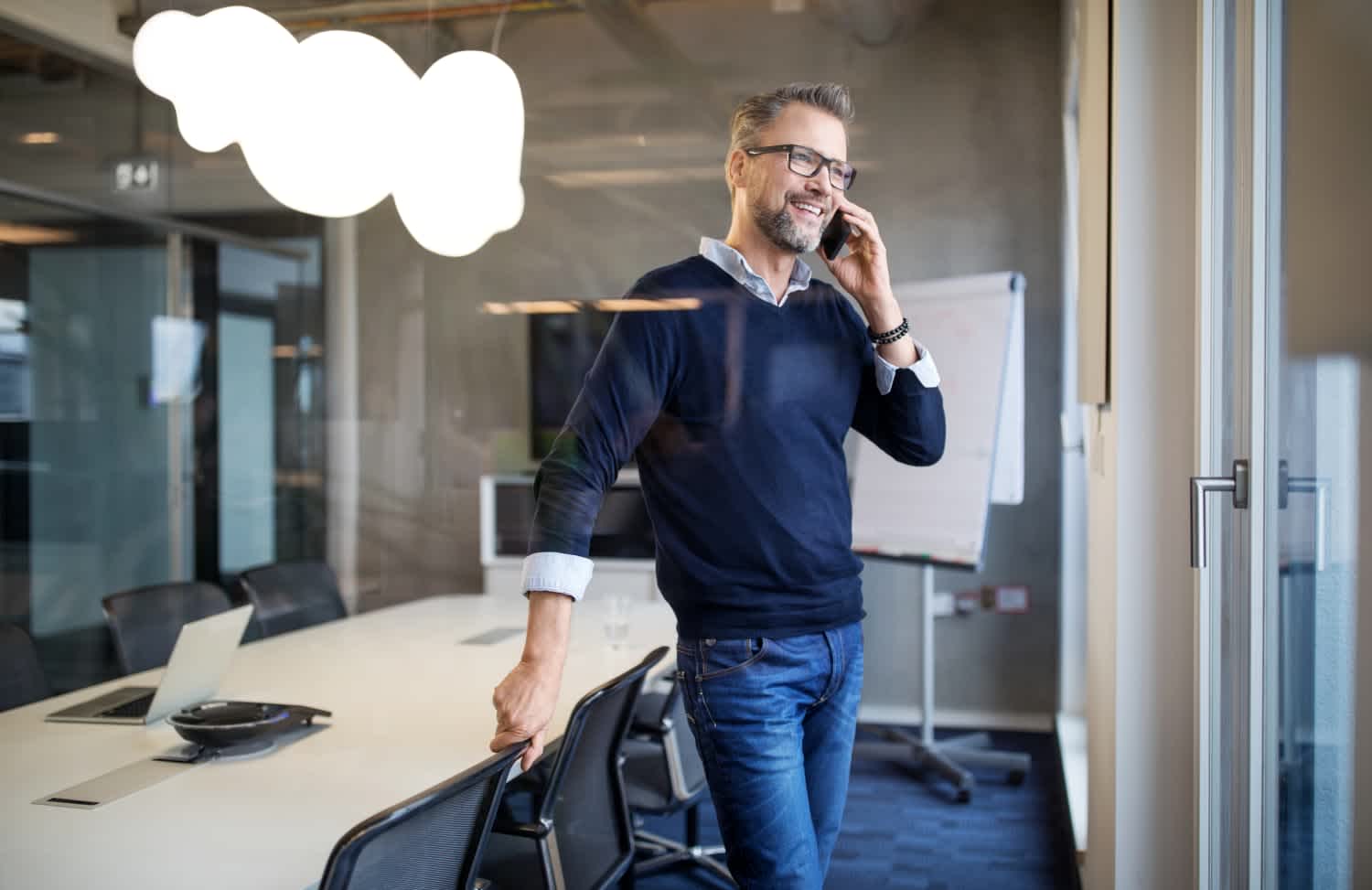 A man wearing glasses and a dark sweater stands in a modern conference room, smiling while talking on a smartphone. He leans on a chair, with a laptop and flipchart visible in the background.
