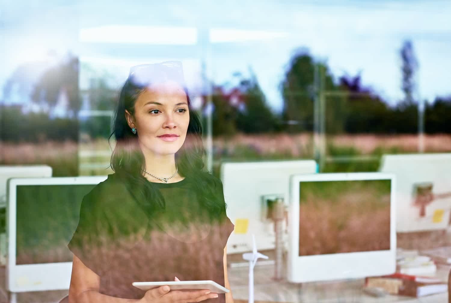 A woman holding a tablet stands behind a glass window, looking outside thoughtfully. The reflection shows trees and grass; behind her are computer monitors in an office setting.