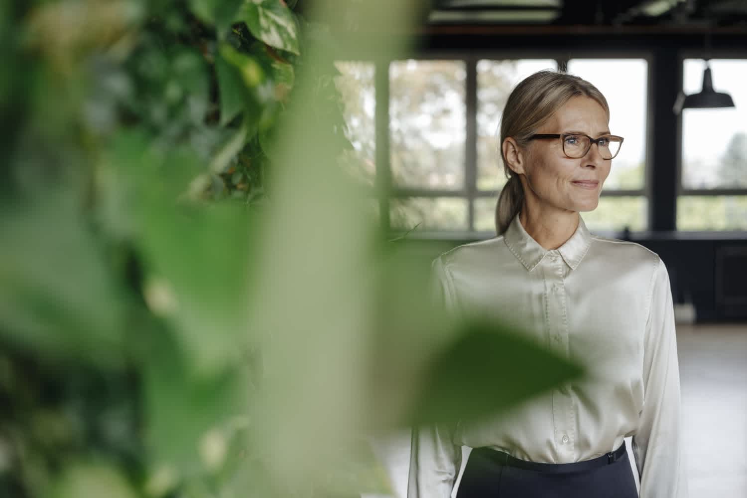 A woman with glasses and a ponytail, wearing a shiny light blouse, stands indoors near large windows and green plants, looking thoughtfully to the side.