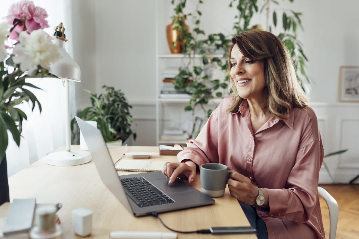 A woman in a pink blouse sits at a desk with a laptop, holding a mug and smiling, surrounded by indoor plants and soft natural light from a nearby window.