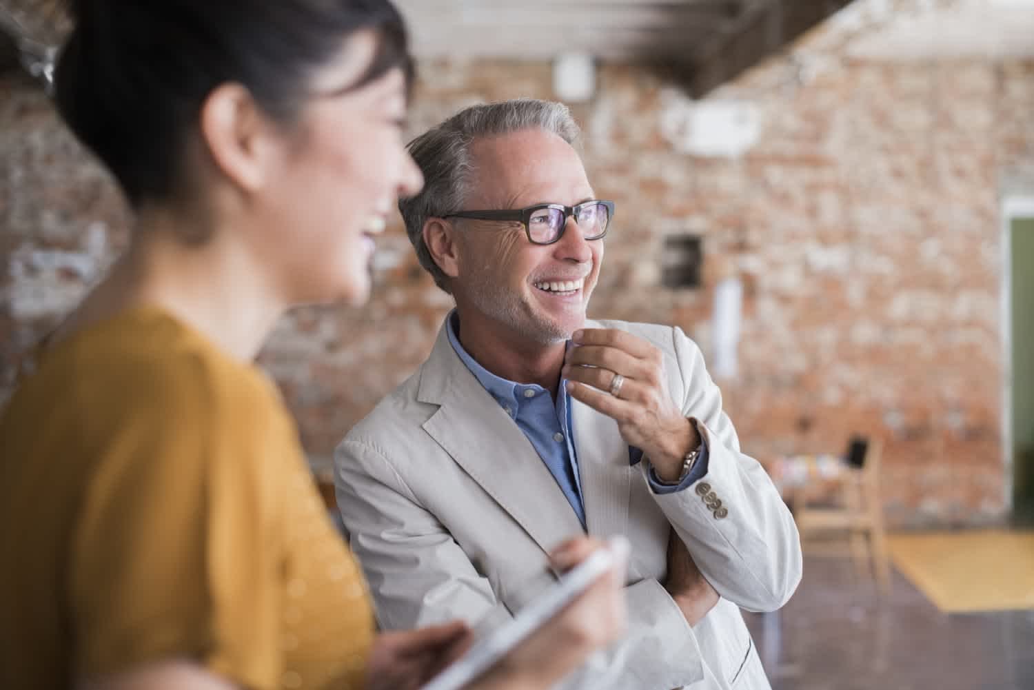 A smiling man in a light suit and glasses stands indoors with his arms crossed, talking to a woman in a yellow top. The background is a blurred brick wall, creating a casual, friendly atmosphere.