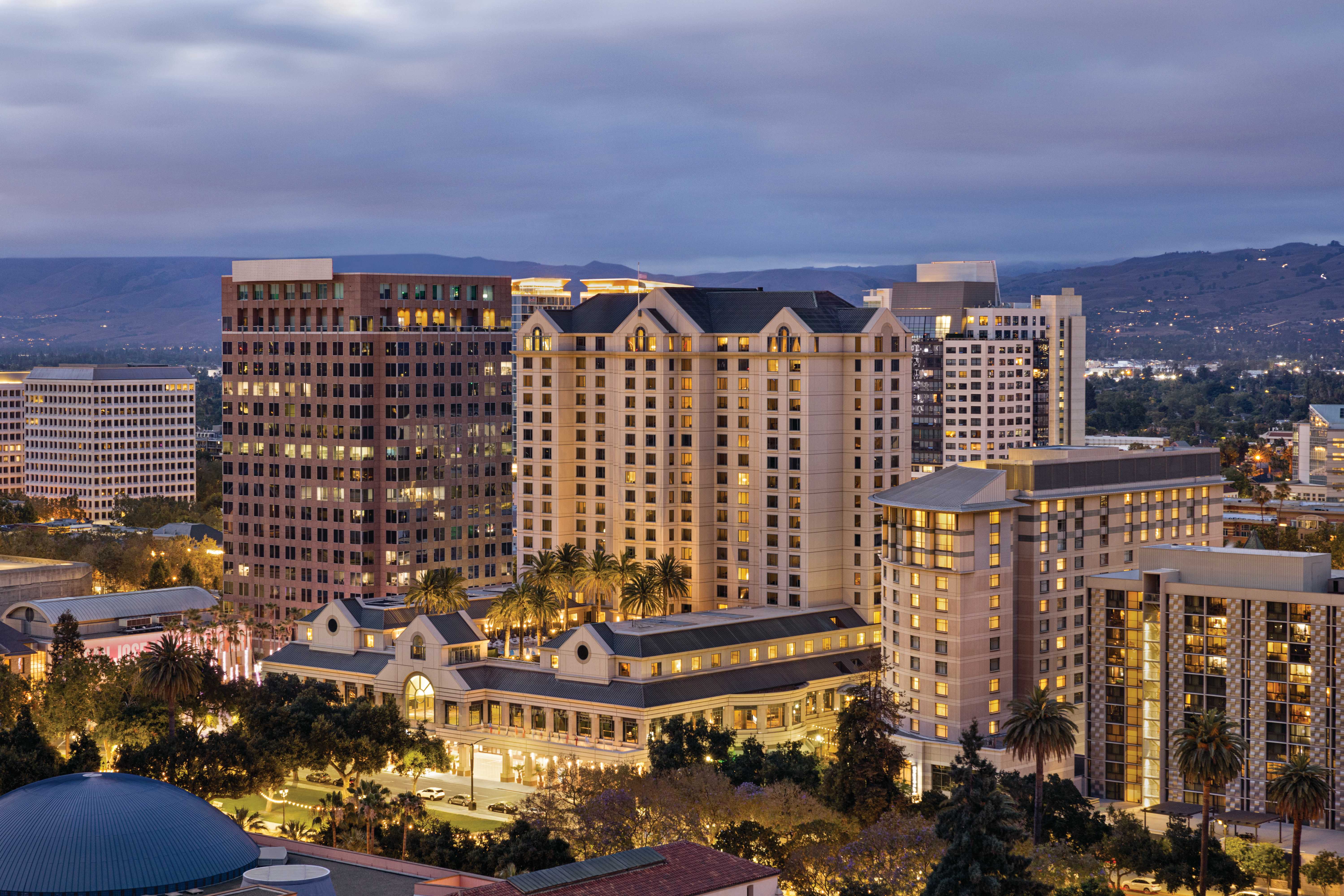 A cityscape at dusk featuring tall buildings with illuminated windows, palm trees, and mountains in the background under a cloudy sky.