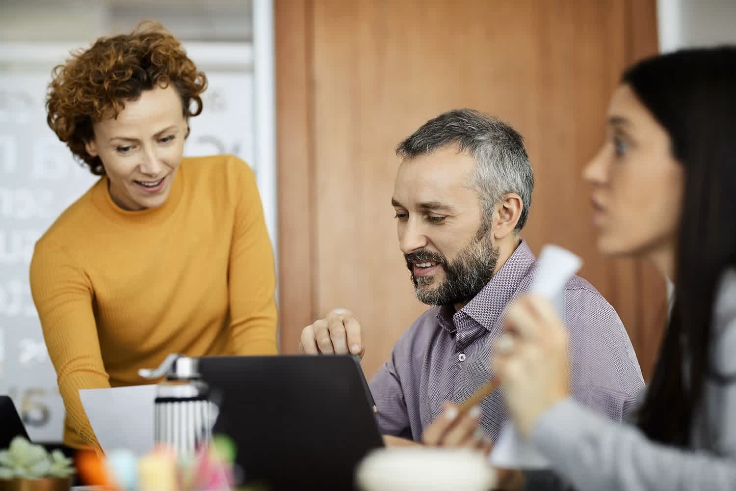 Three people in an office setting work together around a laptop. One person stands smiling and pointing at the screen, while the others sit, one looking at the laptop and one holding papers.