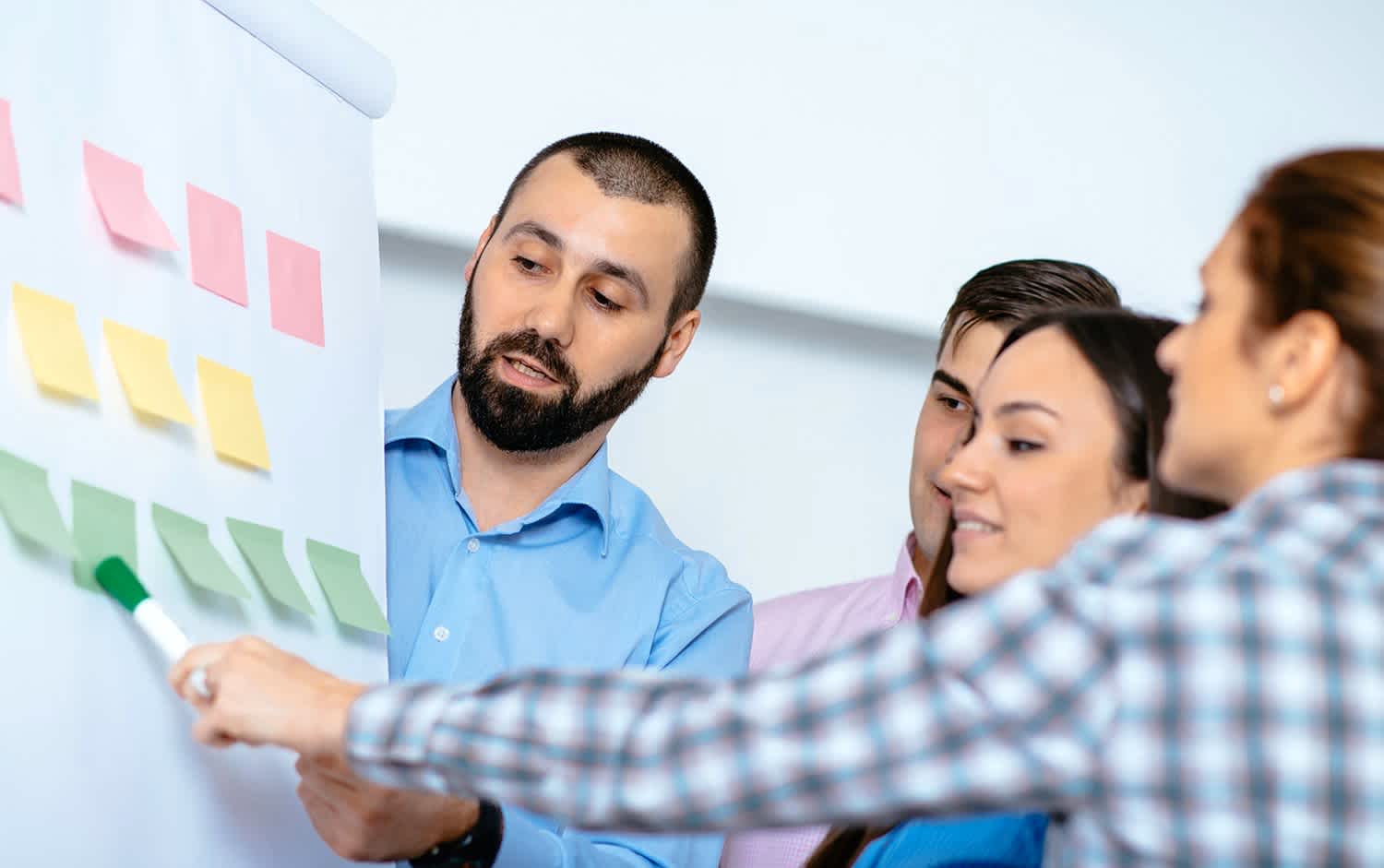Four people stand around a flip chart with colorful sticky notes. One person uses a marker to point at the notes while others attentively look on, suggesting a collaborative discussion or brainstorming session.
