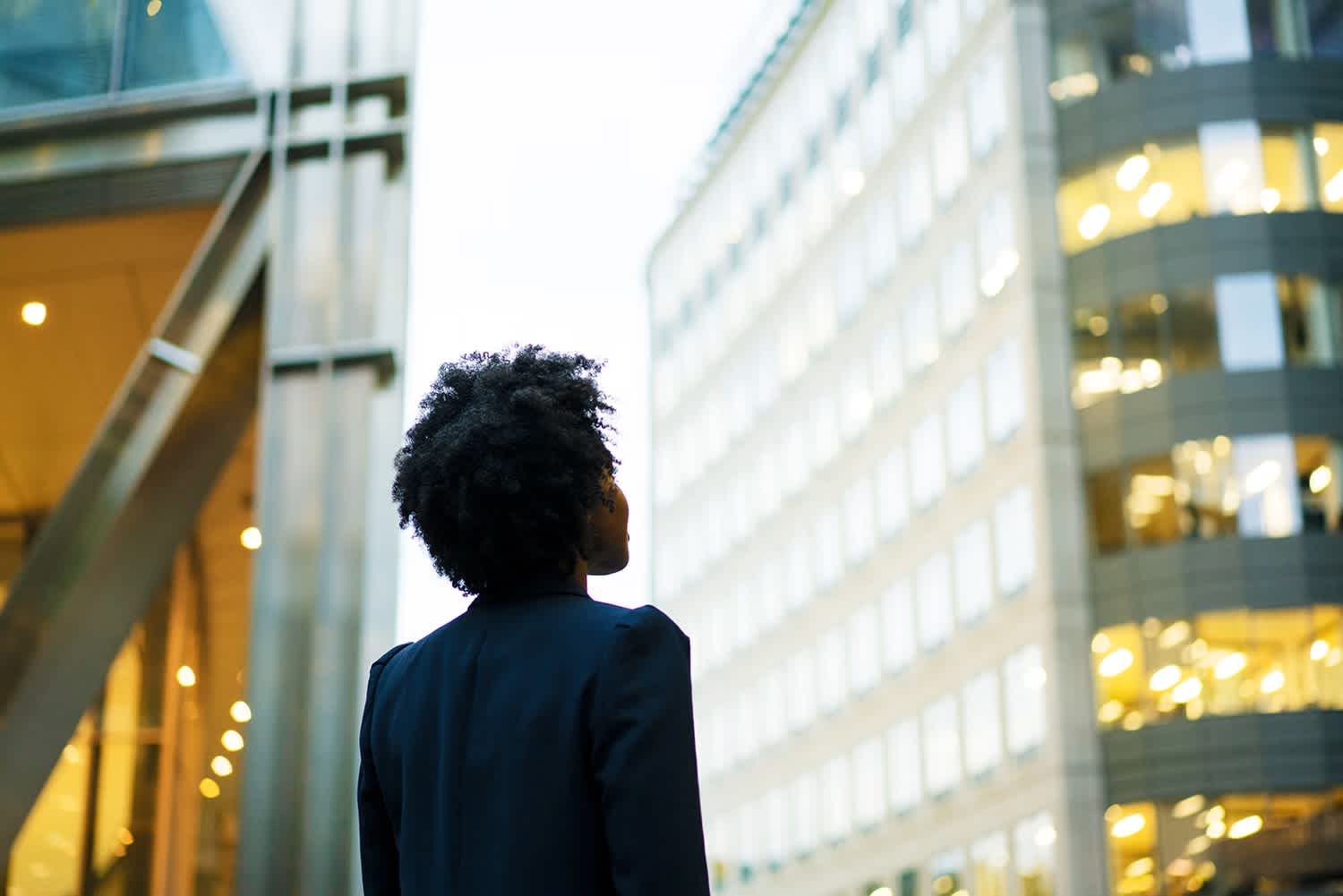 A person with curly hair wearing a dark blazer stands outdoors, looking up at tall, modern office buildings with many windows, some of which are brightly lit. The photo is taken from behind the person.