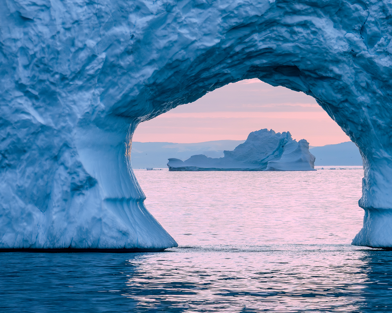 A striking natural ice arch frames a distant iceberg floating on calm Arctic waters. The interior of the ice glows in cool blue tones, contrasting with the soft pink hues of the sunset sky beyond. The smooth water reflects the colors and shapes, creating a serene and almost surreal scene that highlights the beauty and fragility of polar landscapes.