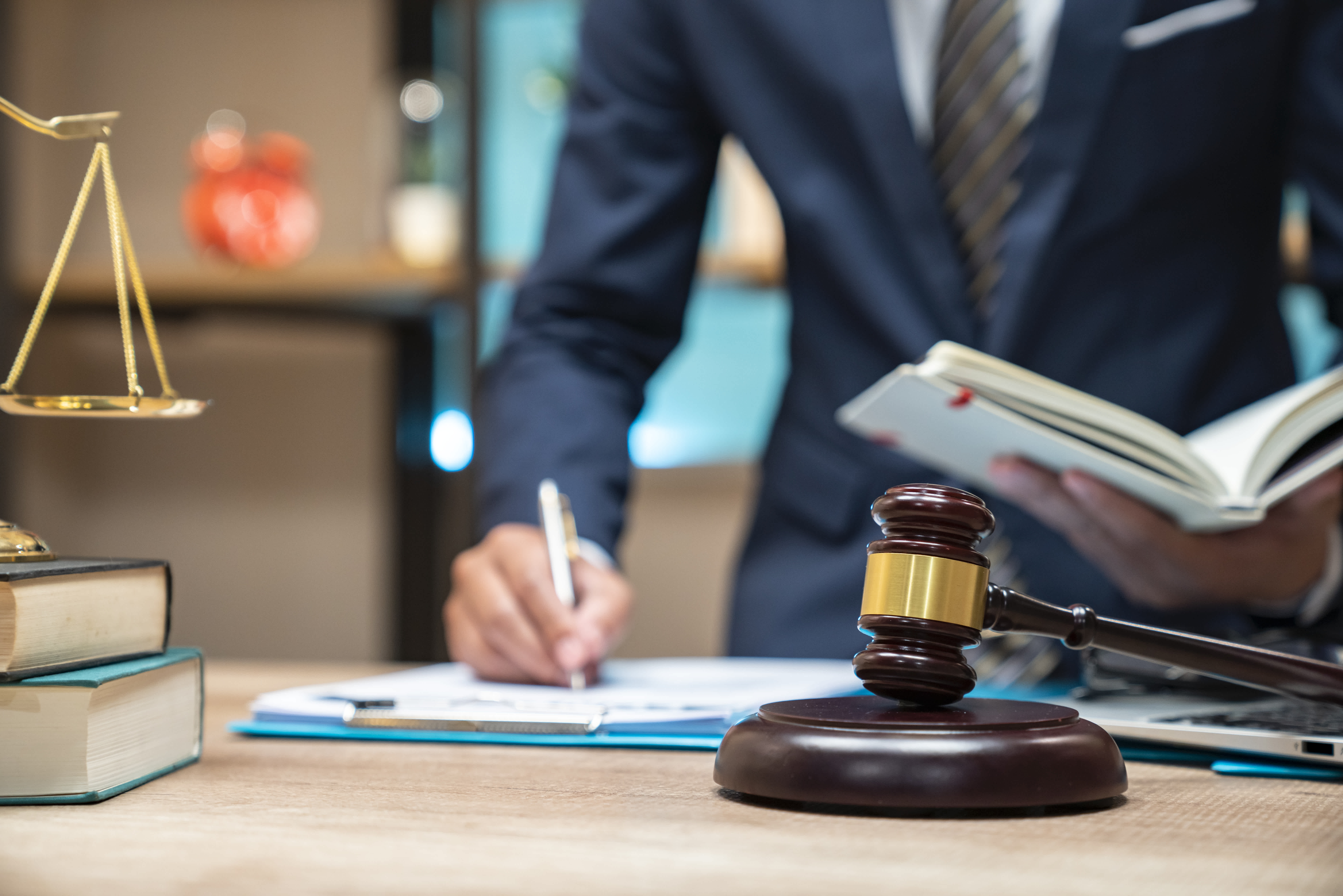 Legal professional signing documents at a desk with a gavel, law books, and scales of justice in the background.