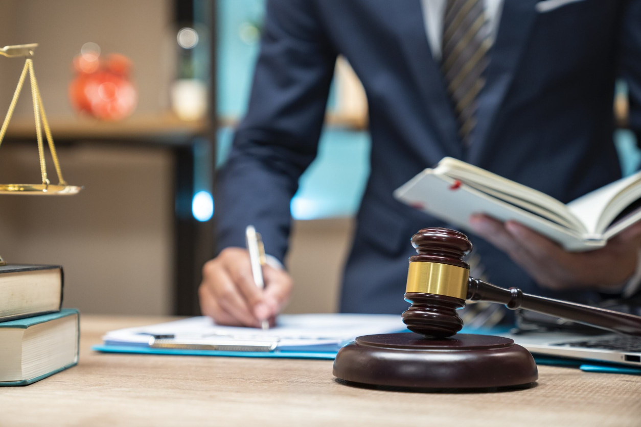 A person in a suit stands at a desk with a gavel, scales of justice, and books, writing in a notebook while holding an open book.
