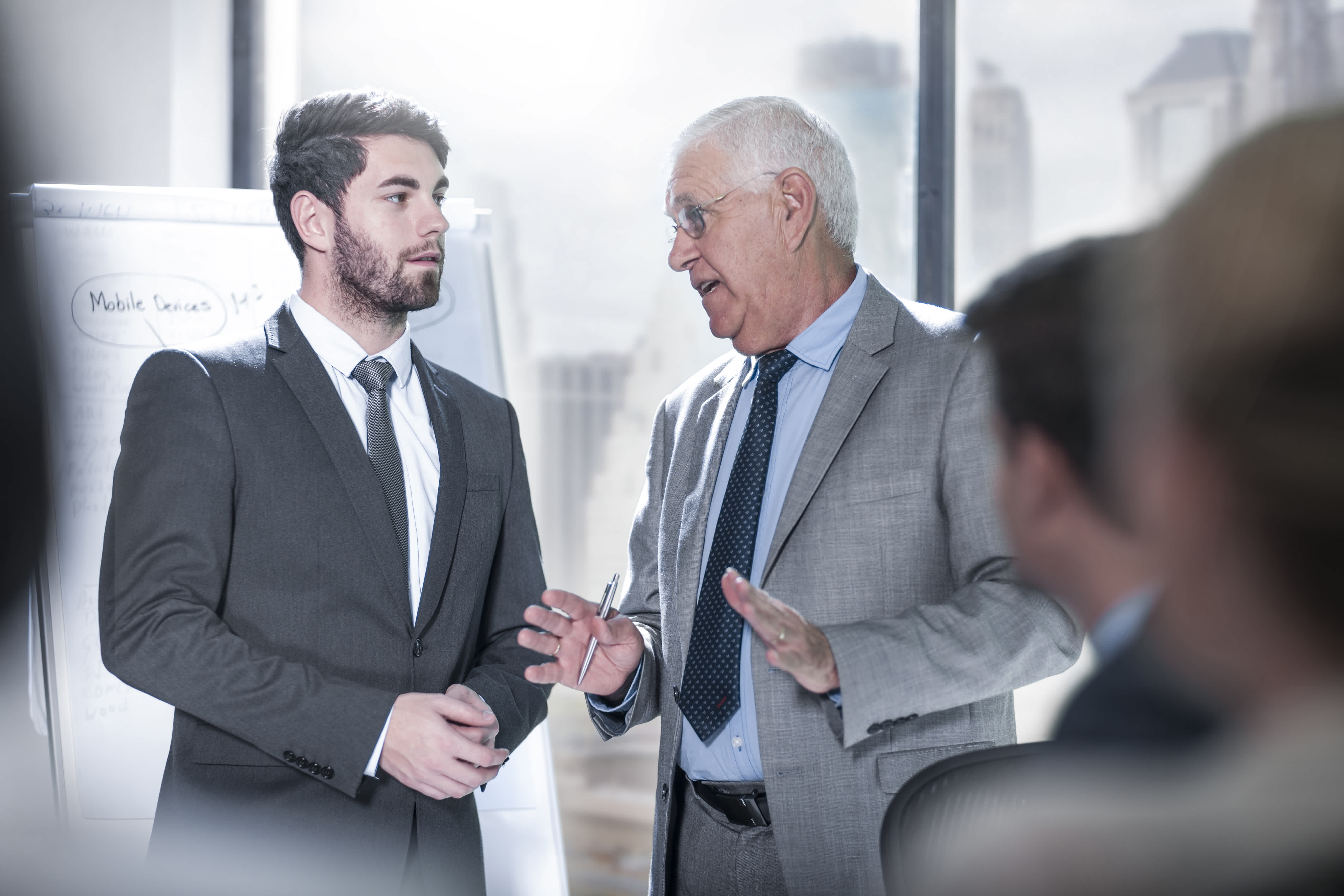Two men in business suits have a serious discussion in an office. One is older with white hair and glasses, gesturing with his hands, while the younger man listens. A flip chart is visible in the background.