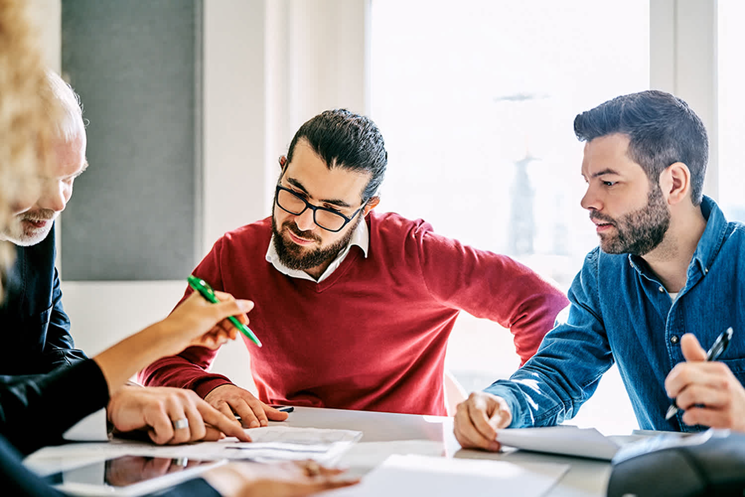 Four people sit around a table engaged in discussion, looking at documents and taking notes. One person is pointing at a paper with a green pen, while the others focus on the materials in front of them.