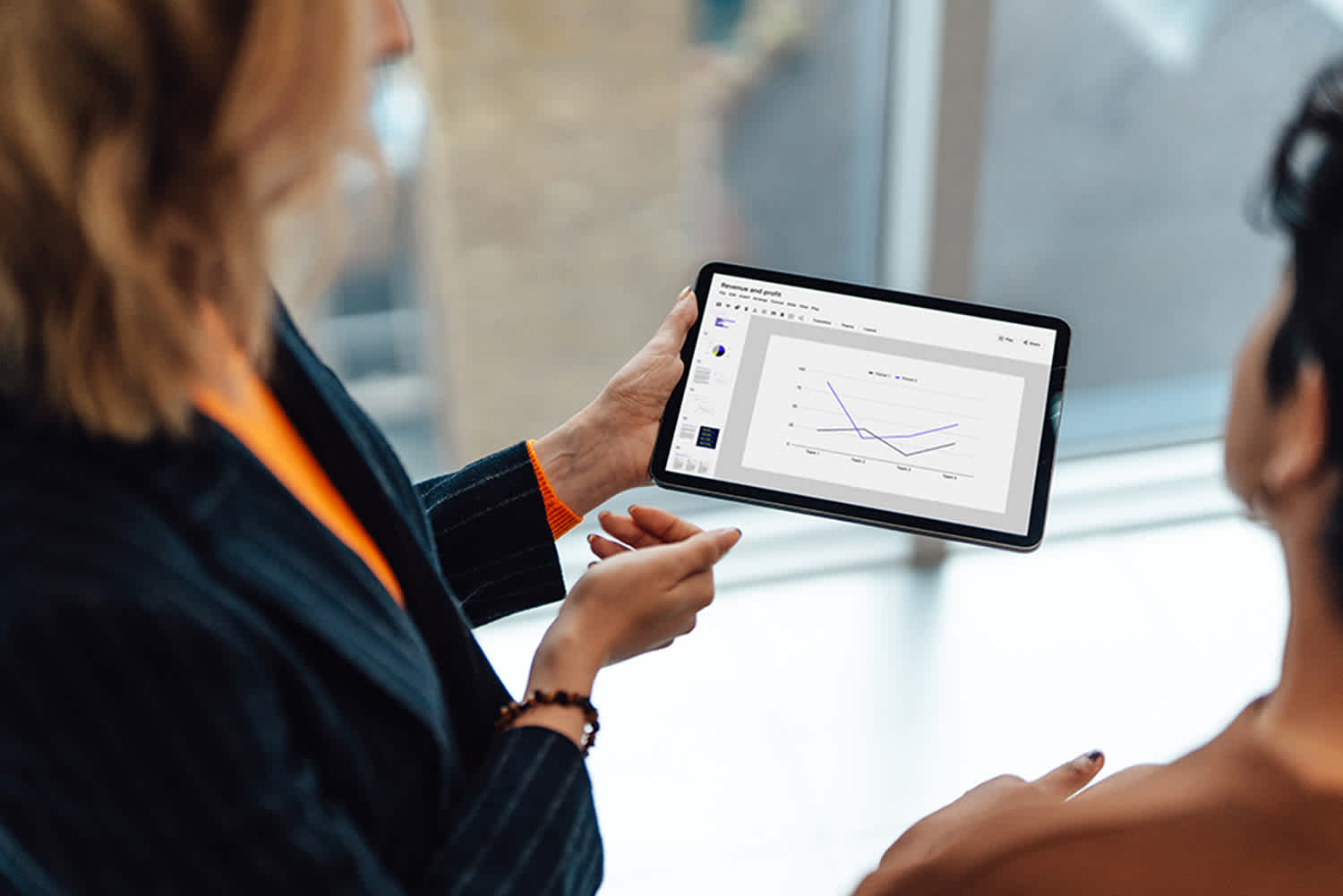 Two people discuss a tablet displaying a line graph with three trend lines, while standing indoors near large windows. One person is holding the device and gesturing, the other listens attentively.