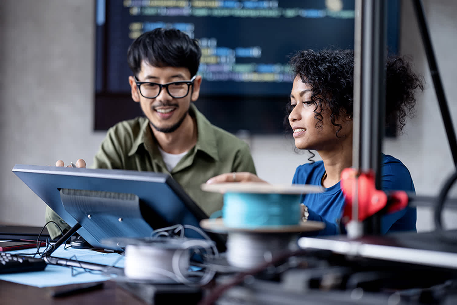 Two people collaborate at a desk with a computer and 3D printer. They work with digital screens and wires, with code visible on a monitor in the background, suggesting a technology or engineering project.
