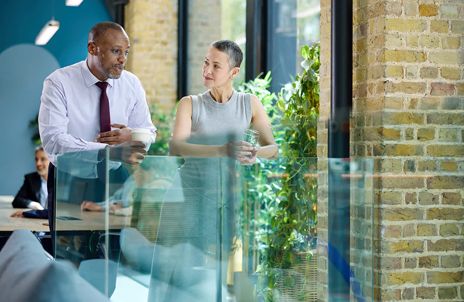 Two colleagues stand by a glass railing in a modern office, engaged in conversation. One holds a coffee cup, the other a glass of water. Large windows and plants create a bright, open atmosphere.