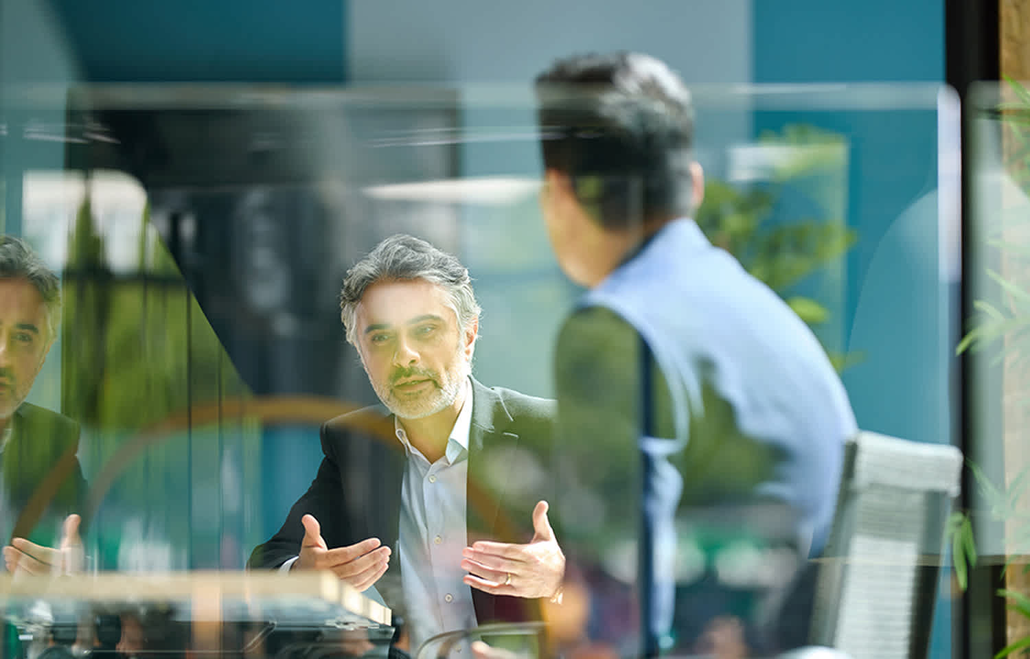 Two men in business attire have a conversation across a table in a modern office setting, separated by a glass partition. One man gestures with his hands while speaking, while the other listens attentively with his back to the camera.