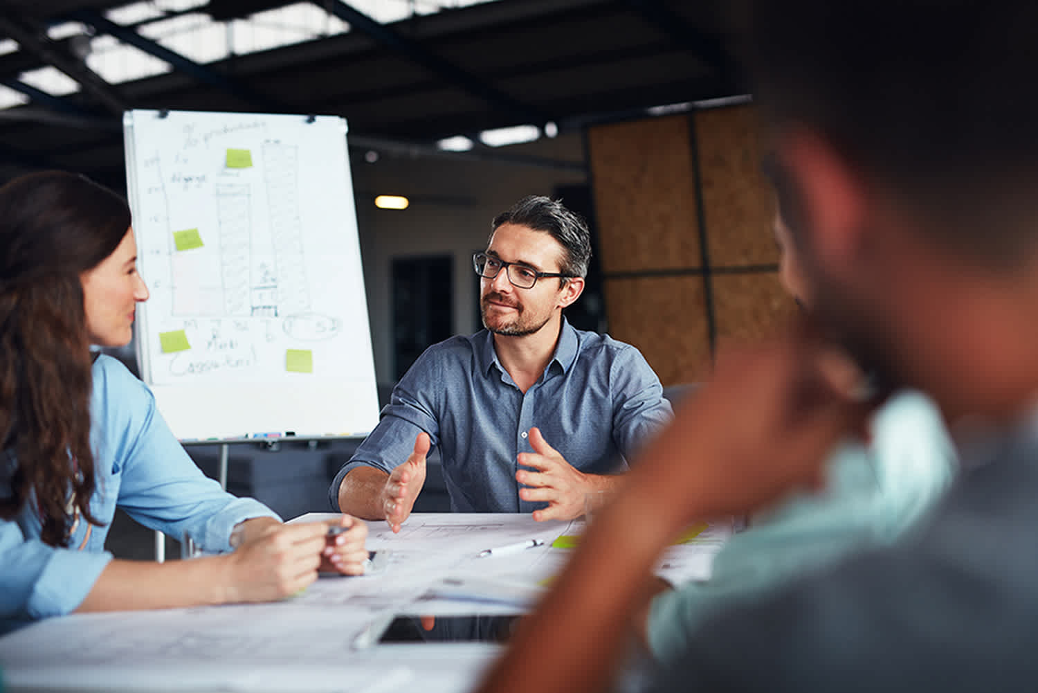 A man in glasses speaks to colleagues during a meeting at a table, with papers and blueprints in front of them. Behind them is a flip chart with graphs and notes in an office setting.