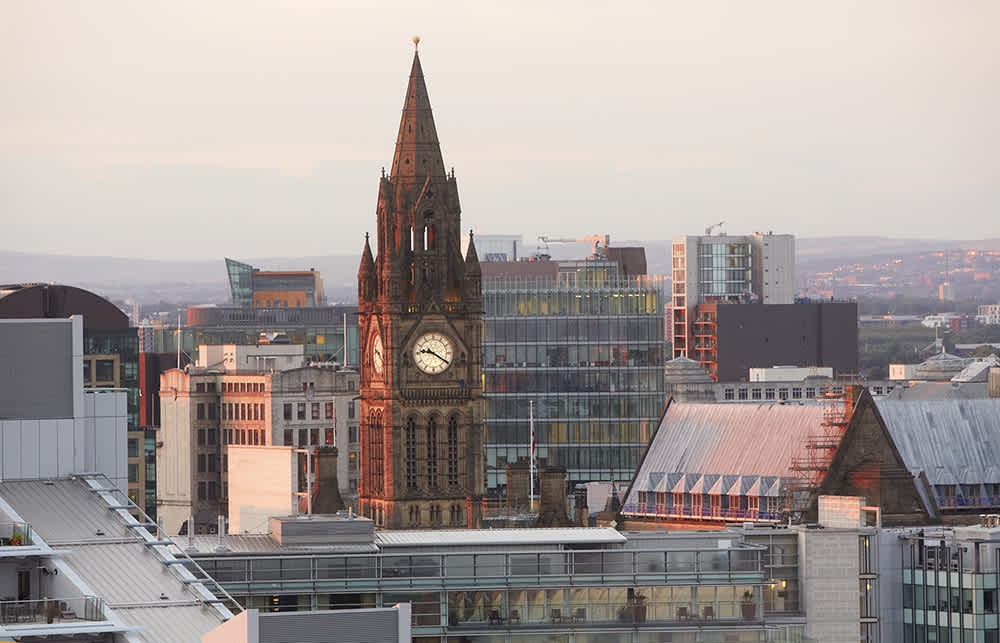 A cityscape at sunset featuring Manchester Town Hall’s clock tower rising above modern and historic buildings, with distant hills in the background.