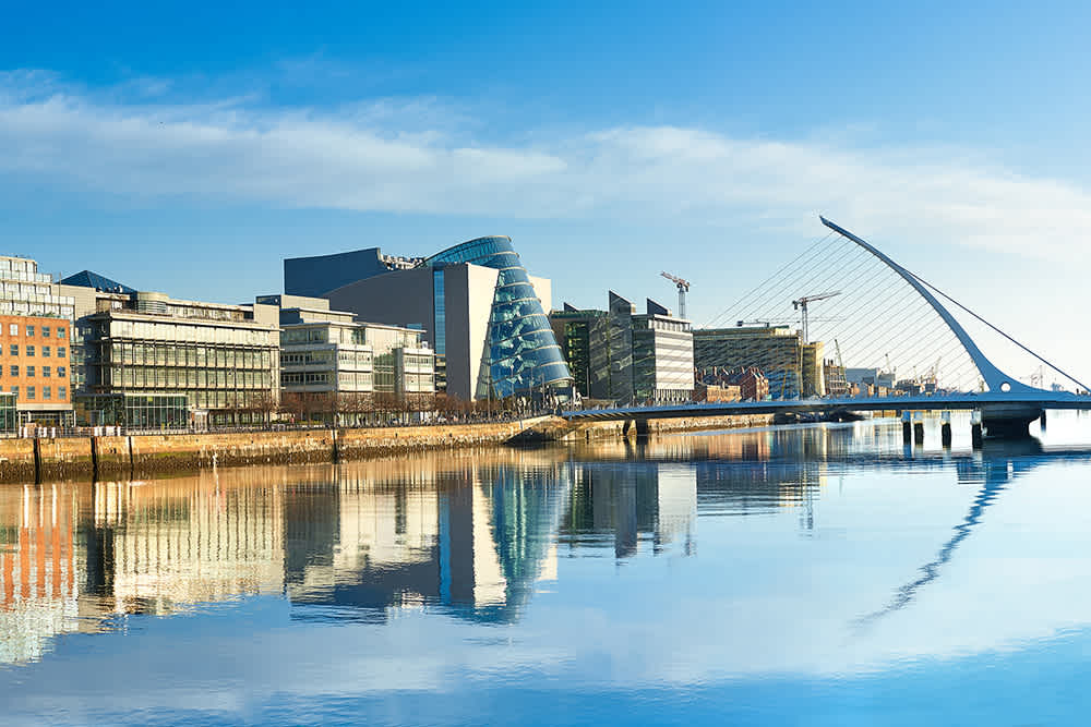 A modern cityscape with glass buildings, including the distinctive curved Convention Centre Dublin, and the Samuel Beckett Bridge over a calm river reflecting the skyline under a clear blue sky.