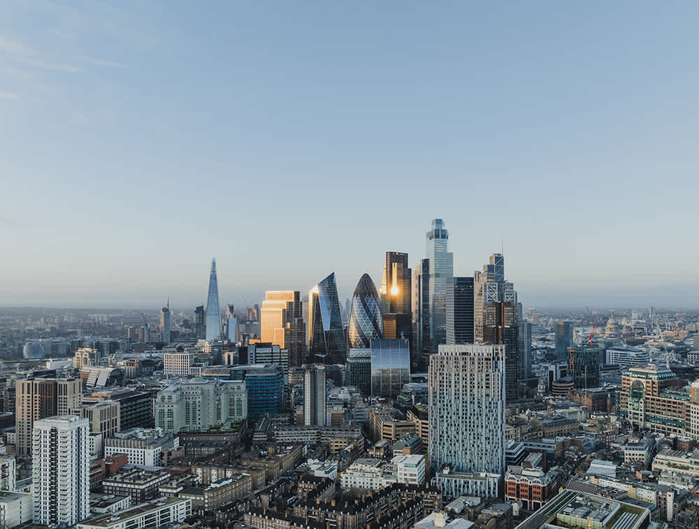 Aerial view of London’s modern skyline at sunrise, featuring iconic skyscrapers like The Shard, The Gherkin, and other glass buildings reflecting sunlight among older city structures.