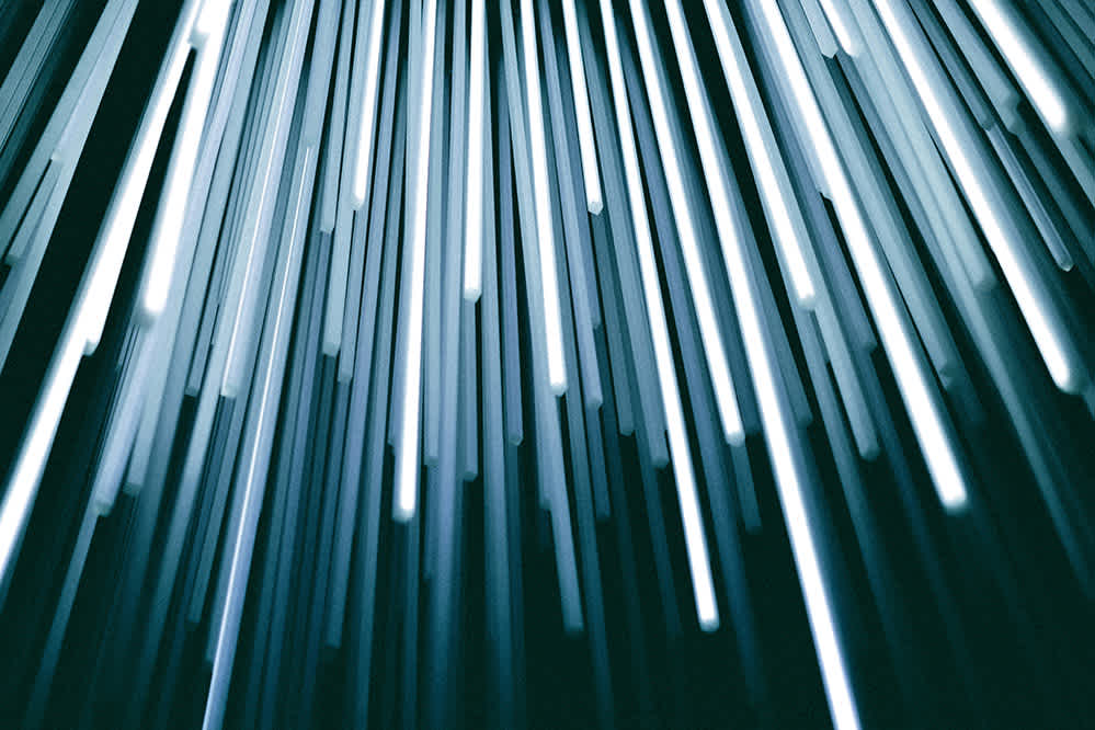 Vertical streaks of white and blue light appear to cascade downward on a dark background, creating an abstract, futuristic pattern resembling fiber optic cables or falling light beams.