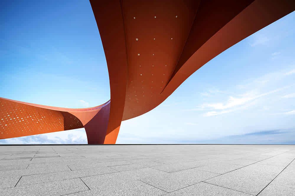 A wide, empty stone plaza faces a dramatic, modern architectural structure with bold, curving orange beams set against a blue sky with scattered clouds.