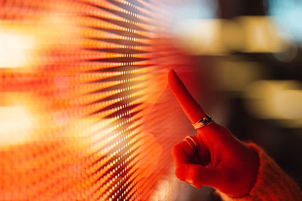 A hand with rings touches a brightly lit, orange and yellow LED screen, causing a ripple effect across the illuminated surface.